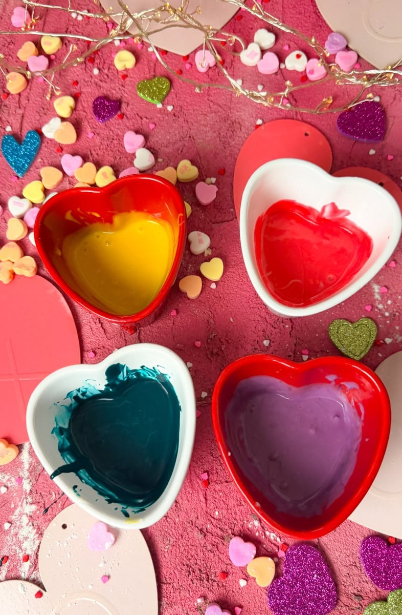 Red, yellow, purple and dark royal icing in heart shaped bowls for decorating heart shaped sugar cookies for Valentine’s Day conversation hearts cookies recipe
