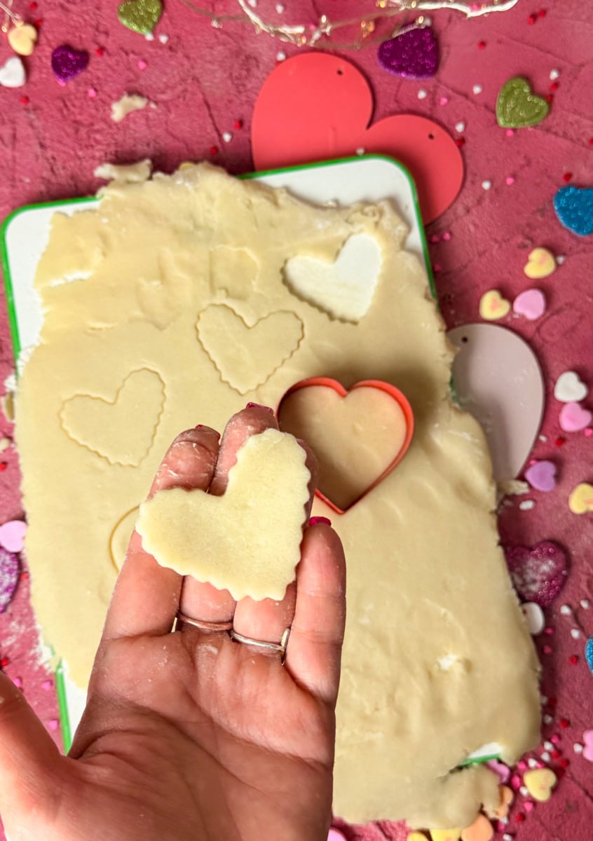Hand holding heart shaped cookie cutouts from rolled sugar cookie dough for Valentine’s Day conversation hearts cookies