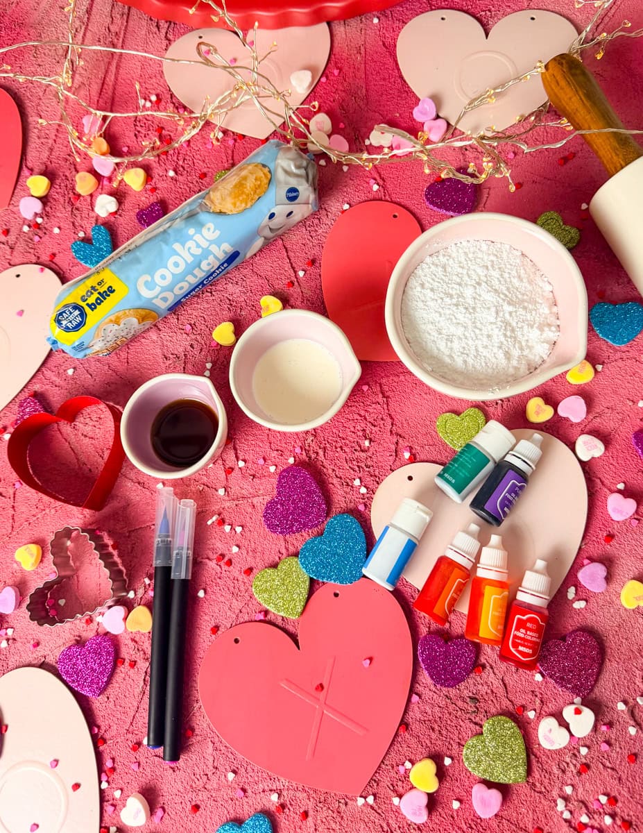 Ingredients for conversation hearts cookies including store-bought sugar cookie dough, powdered sugar, heavy cream, vanilla extract, gel food coloring, edible icing tubes, and a food coloring pen for decorating Valentine’s Day heart-shaped sugar cookies.