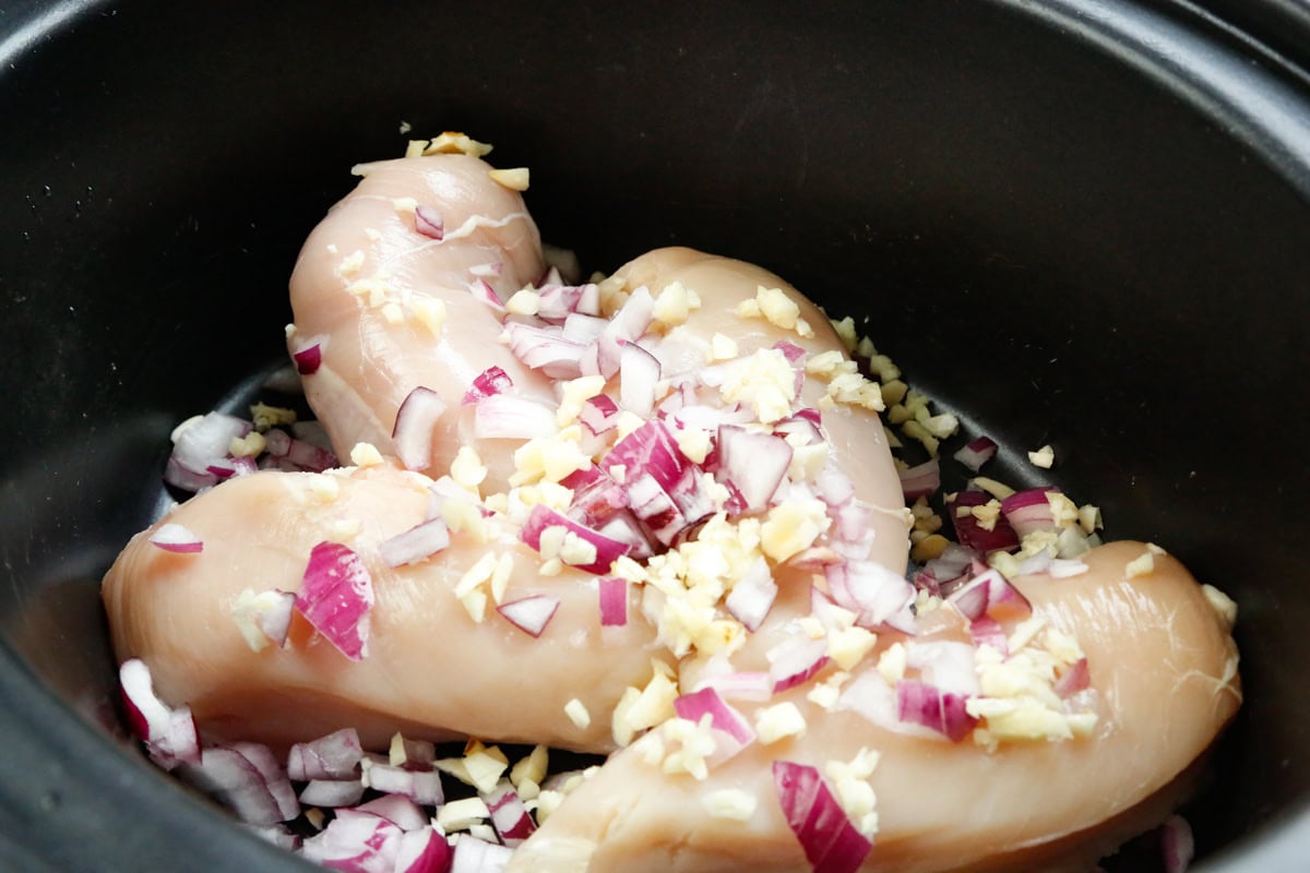 Raw chicken breasts topped with diced red onion and garlic in a slow cooker for BBQ pulled chicken