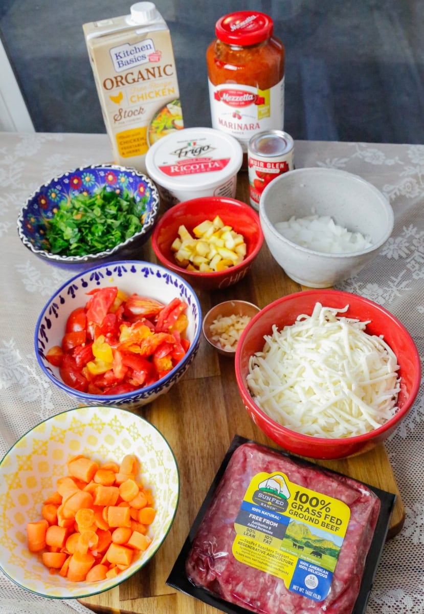 Ingredients for creamy beef ravioli soup including ground beef, carrots, zucchini, spinach, diced tomatoes, mozzarella, ricotta, marinara sauce, tomato paste, onion, garlic, and chicken stock arranged on a wooden board.