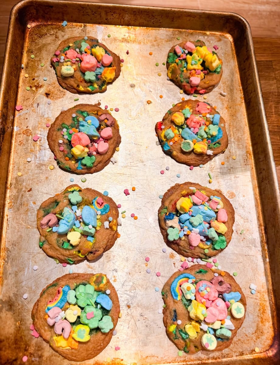 Freshly baked Lucky Charms cookies on a baking sheet topped with marshmallow cereal pieces and colorful sprinkles