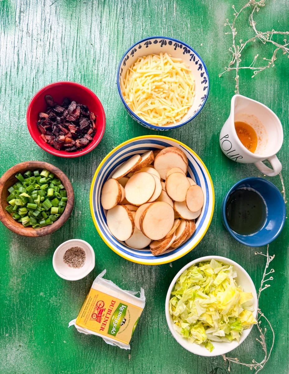 Irish nachos ingredients including sliced russet potatoes, Kerrygold Dubliner cheddar cheese, bacon, chives, shredded lettuce, butter, and seasonings on a green background.