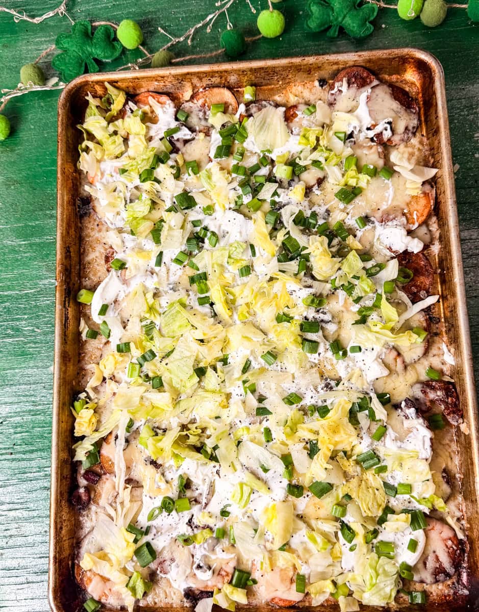 Sheet pan Irish nachos made with crispy roasted potato slices, melted cheese, bacon, sour cream, shredded lettuce, and green onions on a festive St. Patrick’s Day background