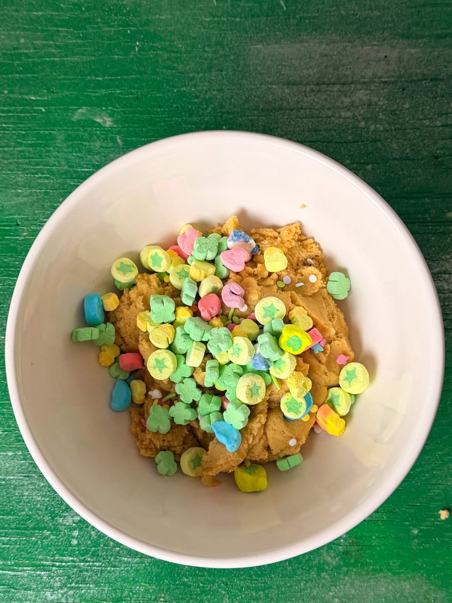 Lucky Charms marshmallows and colorful sprinkles being added to cookie dough in a bowl