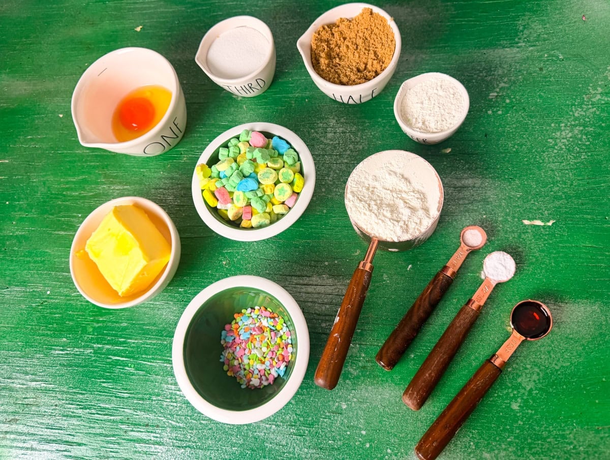 
Ingredients for Lucky Charms cookies including butter, egg, sugar, brown sugar, flour, baking soda, sea salt, vanilla extract, Lucky Charms marshmallows, and sprinkles on a green background