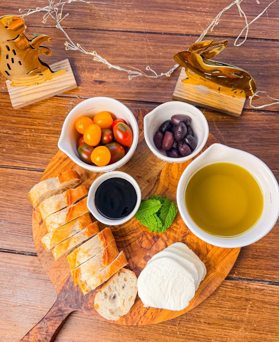 ladybug crostini ingredients including cherry tomatoes mozzarella baguette olives basil leaves and olive oil on wooden board