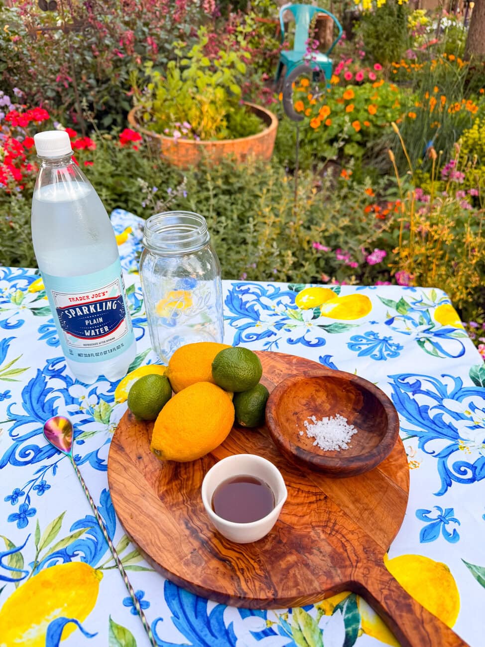 Ingredients for healthy homemade lemon lime soda including sparkling water, fresh lemons, limes, maple syrup, and sea salt arranged on a wooden board in a colorful cottage garden.