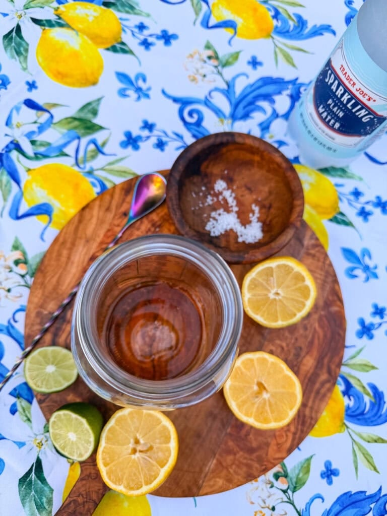 Step one for healthy homemade lemon lime soda recipe showing maple syrup and Baja sea salt added to the bottom of a mason jar with fresh lemon and lime slices nearby.