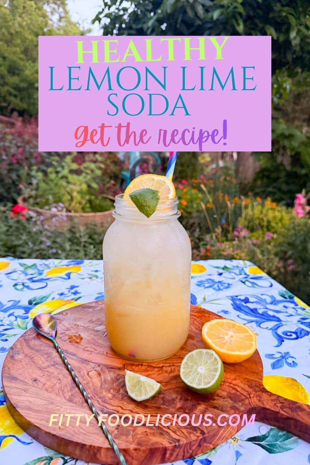 Homemade lemon lime soda served in a mason jar over ice with fresh lemon and lime slices, photographed outdoors in a colorful cottage garden setting with fresh citrus and sparkling summer drink ingredients.