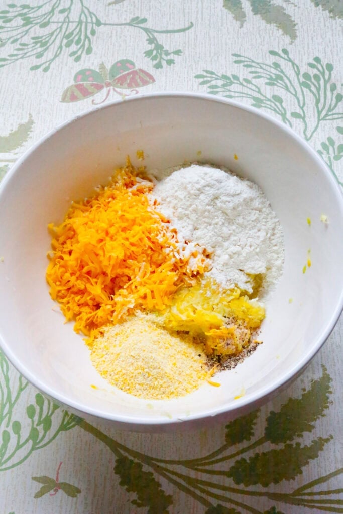 Yellow squash, cheddar cheese, cornmeal, flour, and seasonings in a bowl before mixing homemade squash fritters.