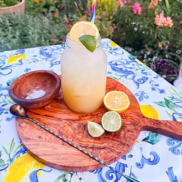Fresh homemade lemon lime soda served over ice in a mason jar with lime garnish, photographed outdoors on a colorful summer table with fresh citrus and a garden background.