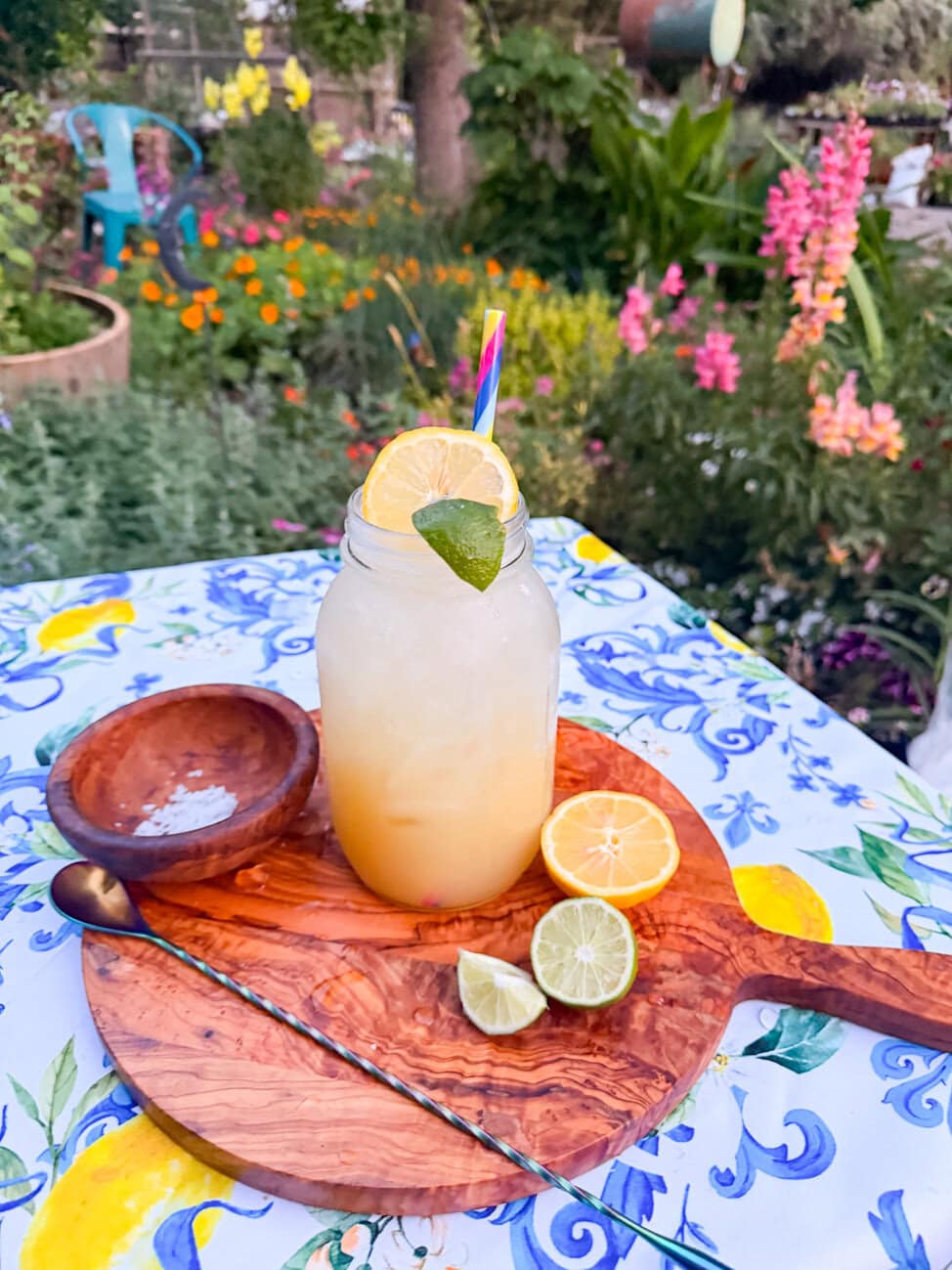 Fresh homemade lemon lime soda served over ice in a mason jar with lime garnish, photographed outdoors on a colorful summer table with fresh citrus and a garden background.