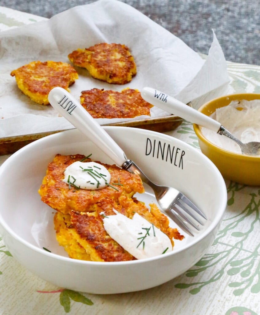 Crispy yellow squash fritters with sour cream and fresh chives served in a white bowl with additional fritters in the background.