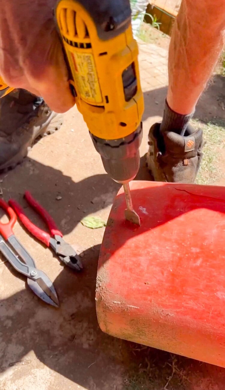 Cordless drill creating a hole in an orange utility bucket for a DIY outdoor hose and extension cord storage tutorial.