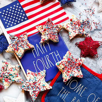 Star-shaped 4th of July Rice Krispie treats with patriotic red, white, and blue sprinkles arranged on a festive Fourth of July display with an American flag.