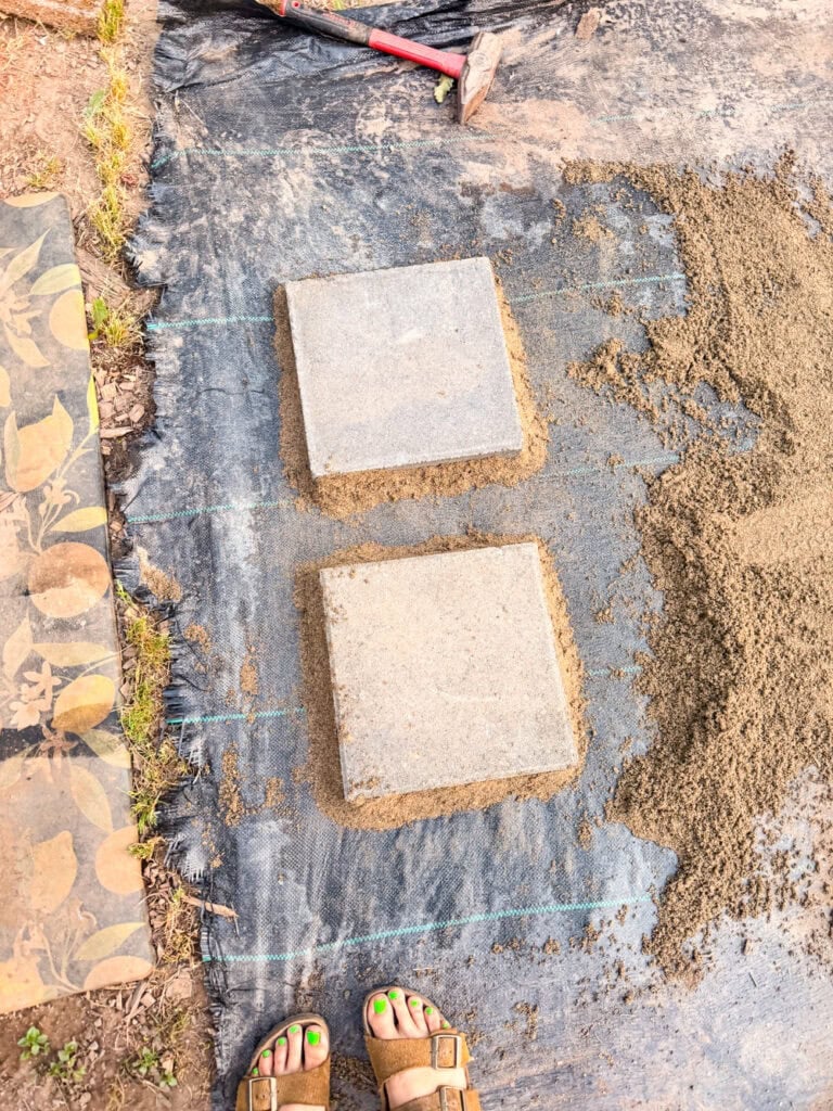 Installing stepping stone pavers on a backyard garden pathway using leveling sand and weed barrier fabric.