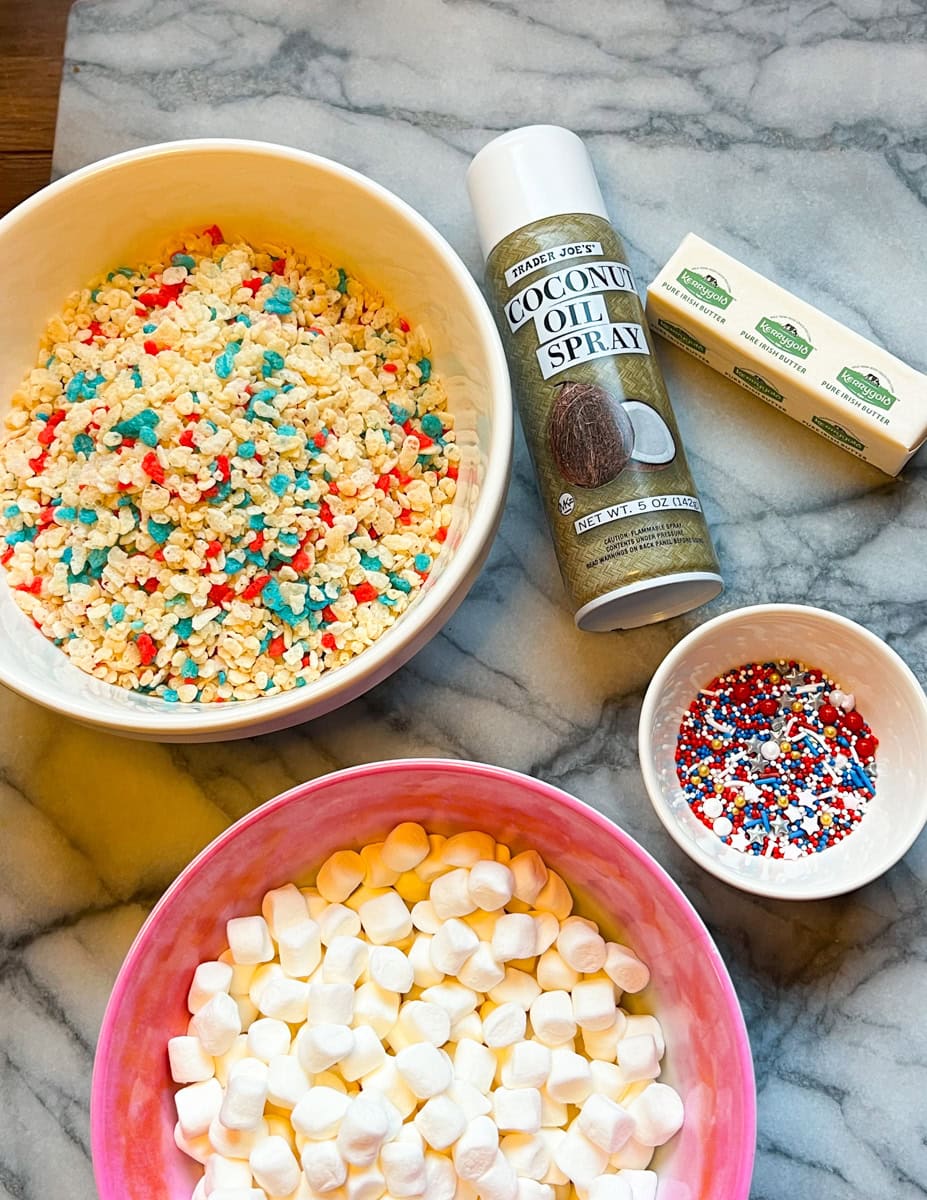 Ingredients for Fourth of July Rice Krispie treats, including marshmallows, butter, vanilla extract, patriotic sprinkles, and star-shaped sprinkles on a marble countertop.