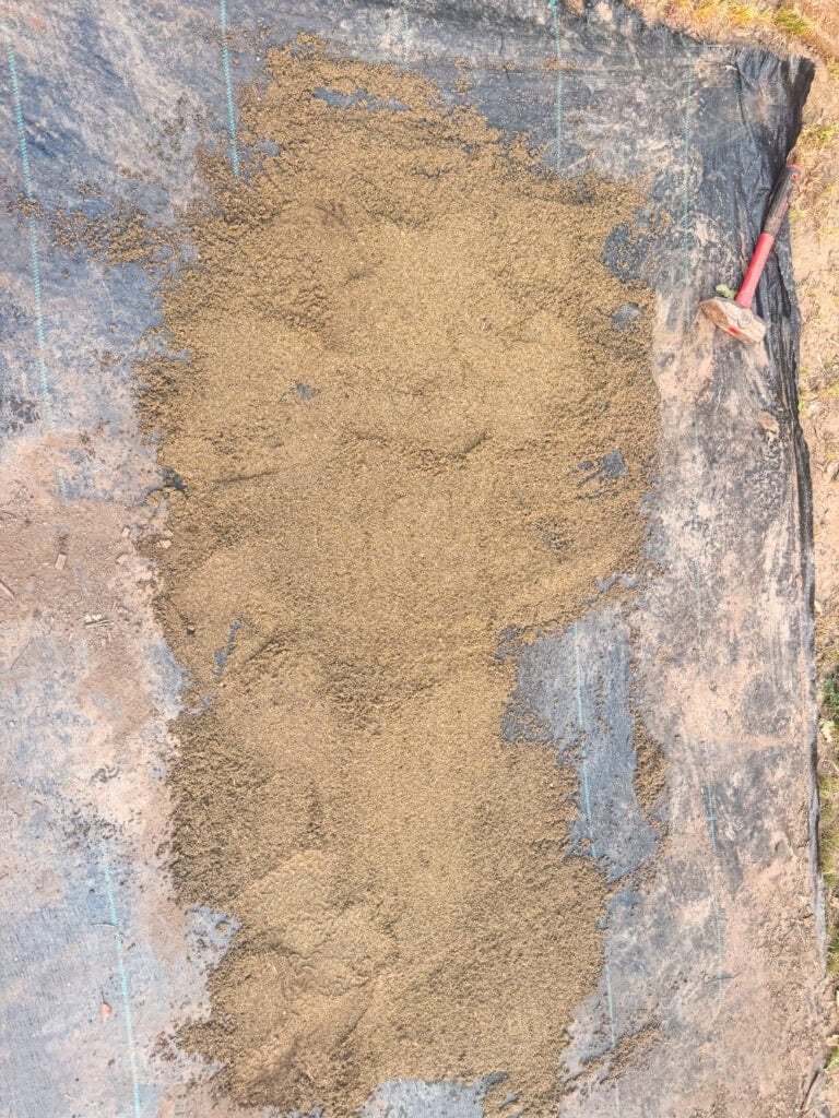 Leveling sand spread over weed barrier fabric to create a stable base for a DIY garden path.