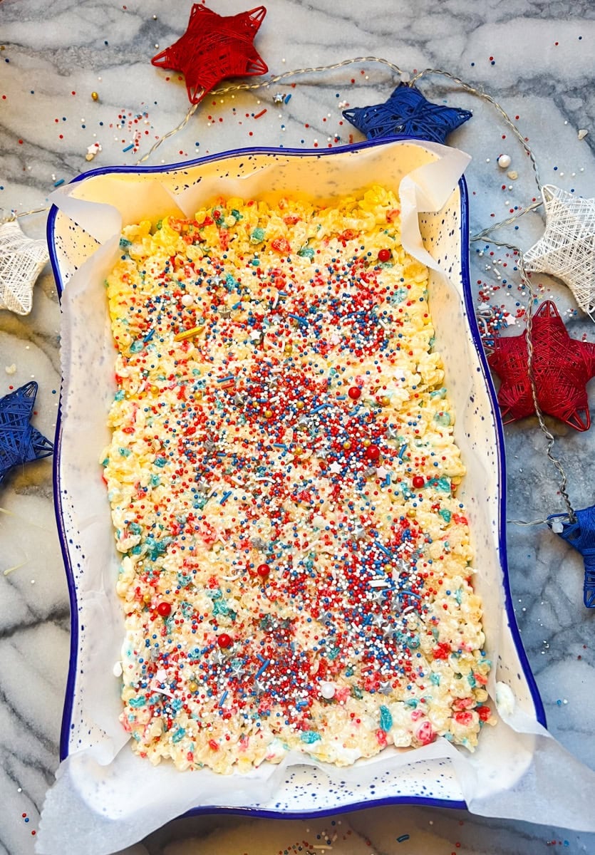 Fourth of July Rice Krispie treats pressed into a parchment-lined baking dish and topped with patriotic red, white, and blue jimmies and star sprinkles.