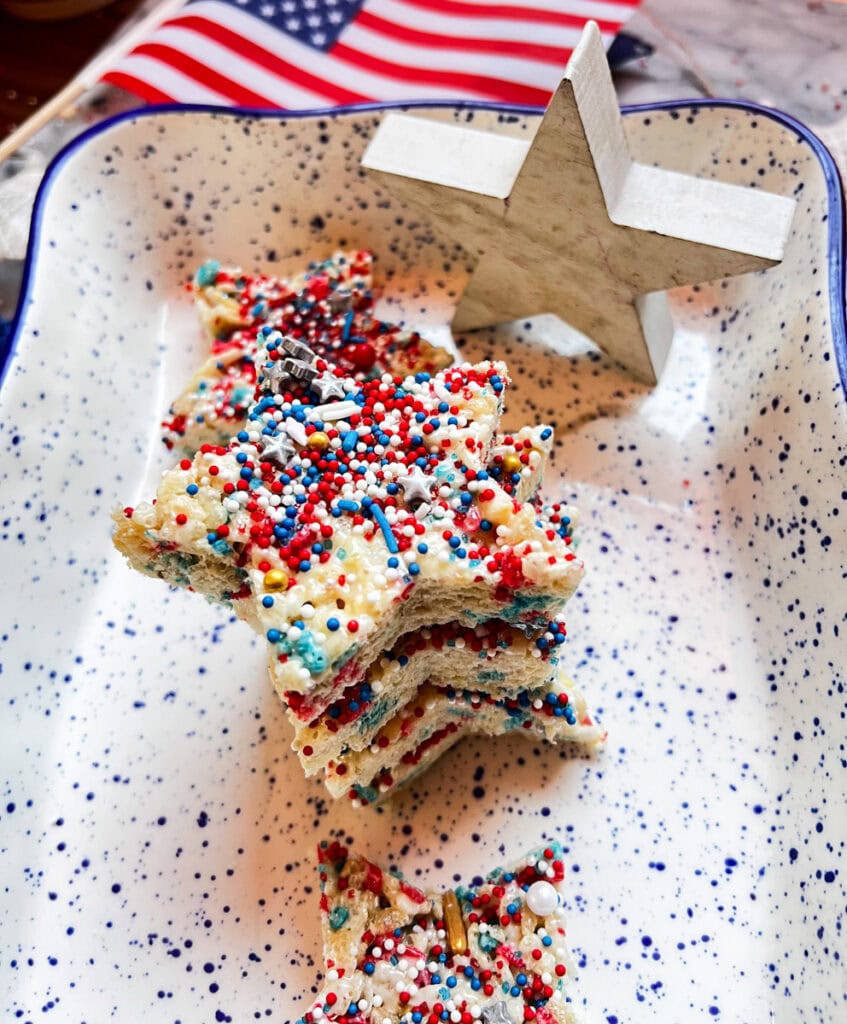 Homemade star-shaped Rice Krispie treats topped with red, white, and blue sprinkles on a white serving platter, perfect for a Fourth of July celebration.