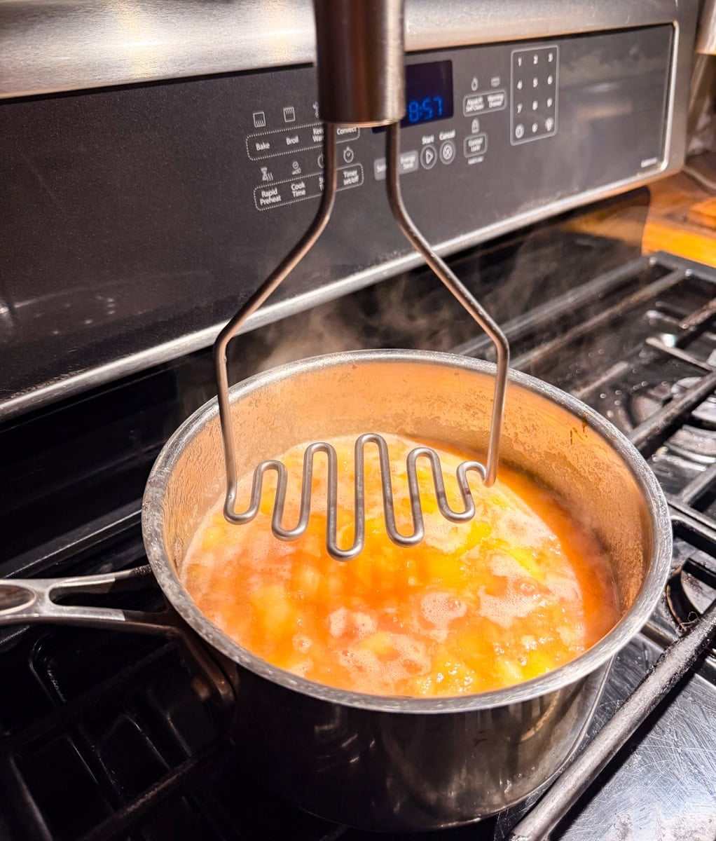 Potato masher breaking down simmering peaches in a saucepan while making fresh peach jam.
