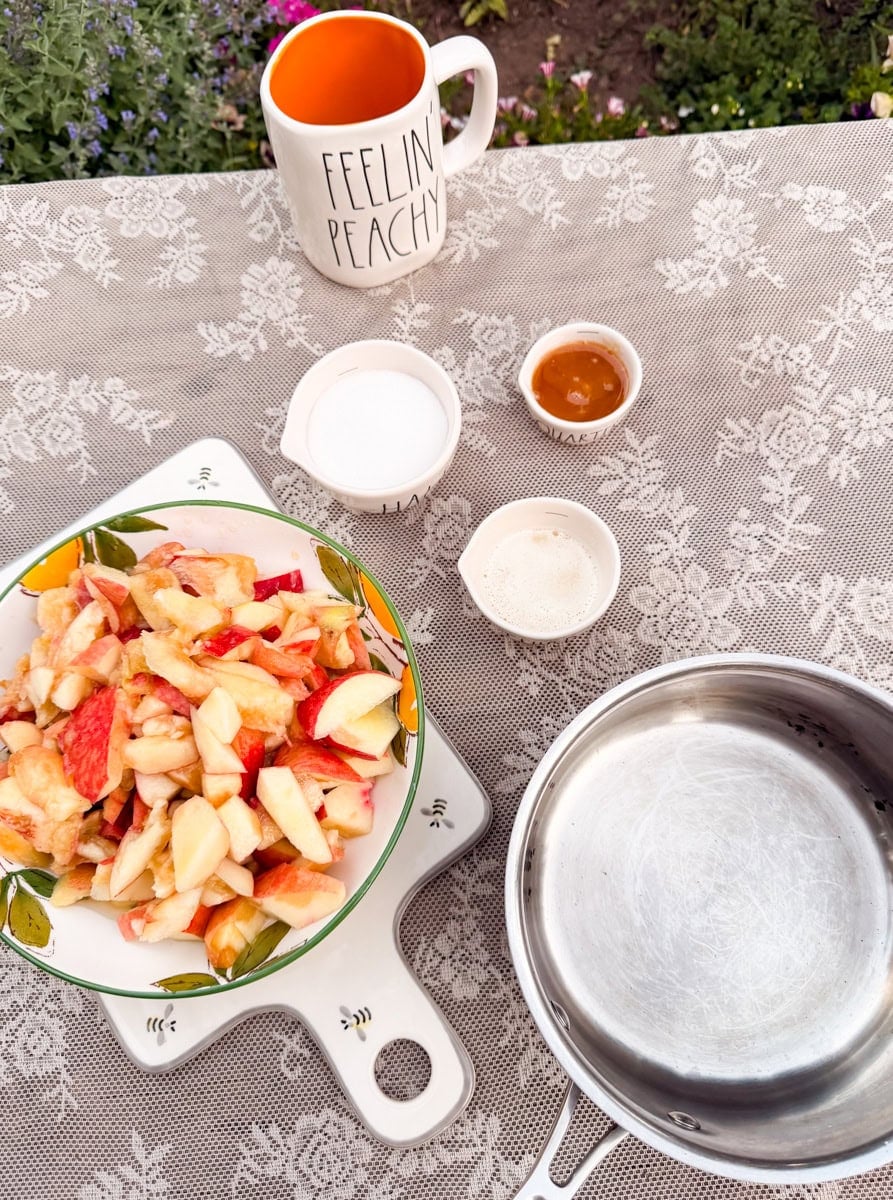 Chopped fresh peaches, honey, sugar, and lemon juice measured beside a saucepan before making peach jam.