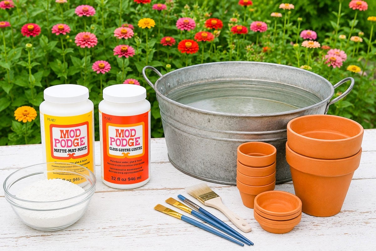 Supplies for aging terracotta pots including terracotta pots, Mod Podge, flour, paint brushes, and a galvanized tub of water on a white table outdoors.
