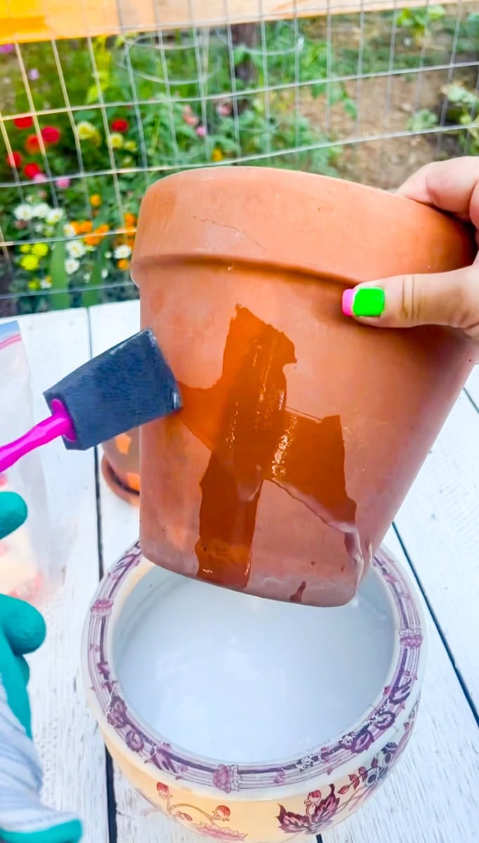Brushing a diluted Mod Podge mixture onto the outside of a terracotta pot to begin creating a weathered, aged appearance.