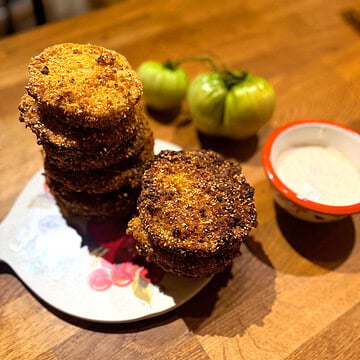 Stacked crispy fried green tomatoes served with dipping sauce and fresh green tomatoes in the background.