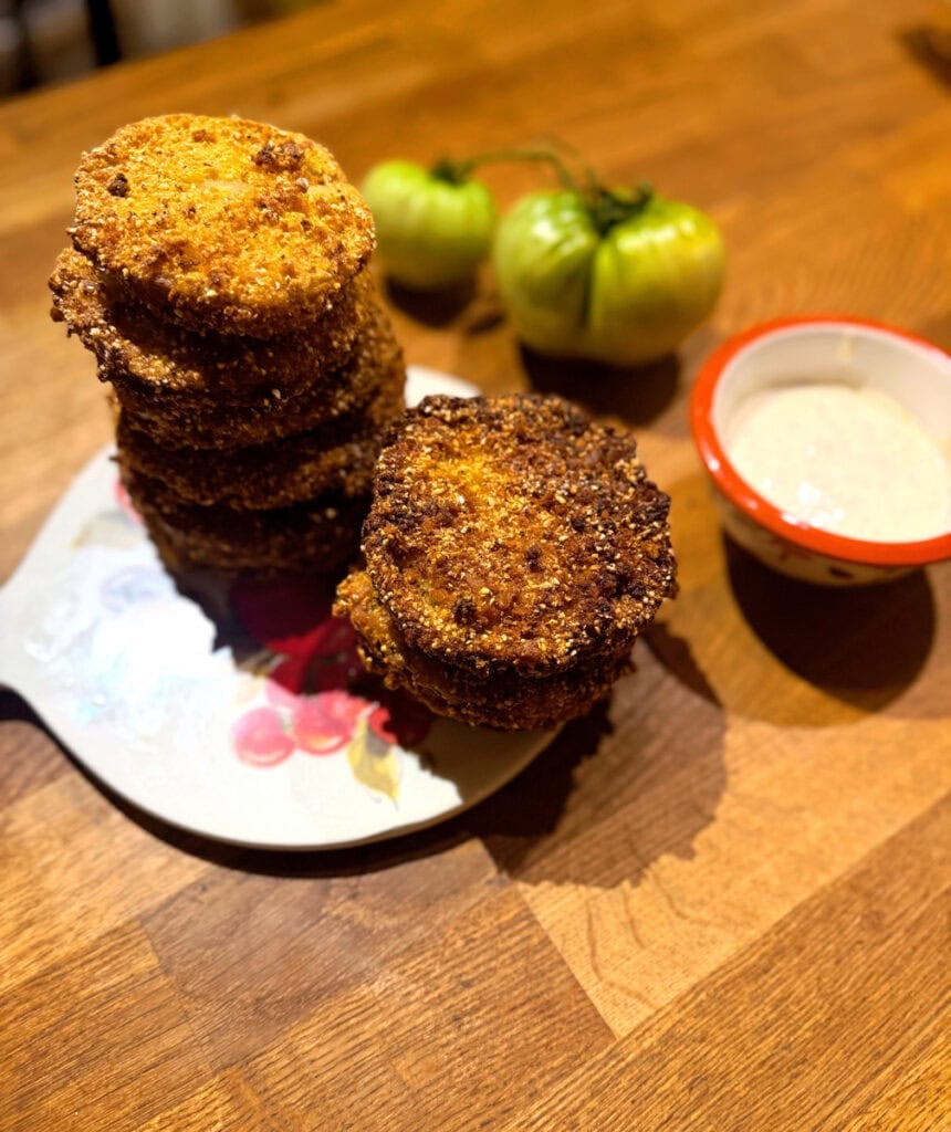Stacked crispy fried green tomatoes served with dipping sauce and fresh green tomatoes in the background.