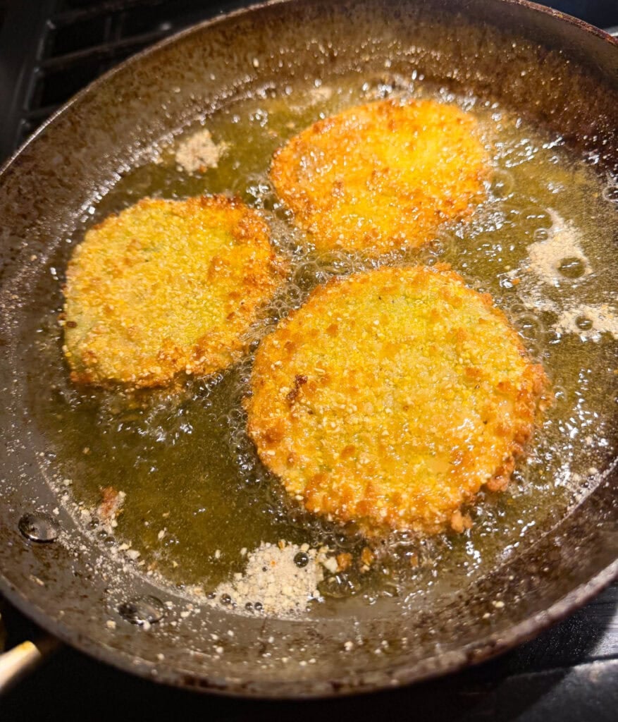 Breaded green tomato slices frying in extra-virgin olive oil in a skillet until golden and crispy.