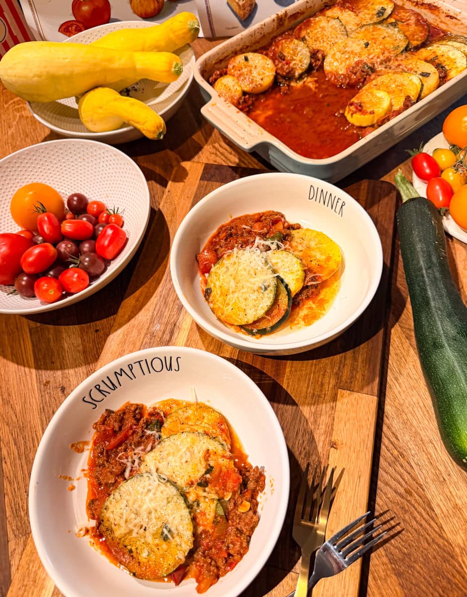 Two bowls of homemade ratatouille served with roasted zucchini, yellow squash, tomatoes, and Parmesan cheese alongside the baking dish and fresh garden vegetables.