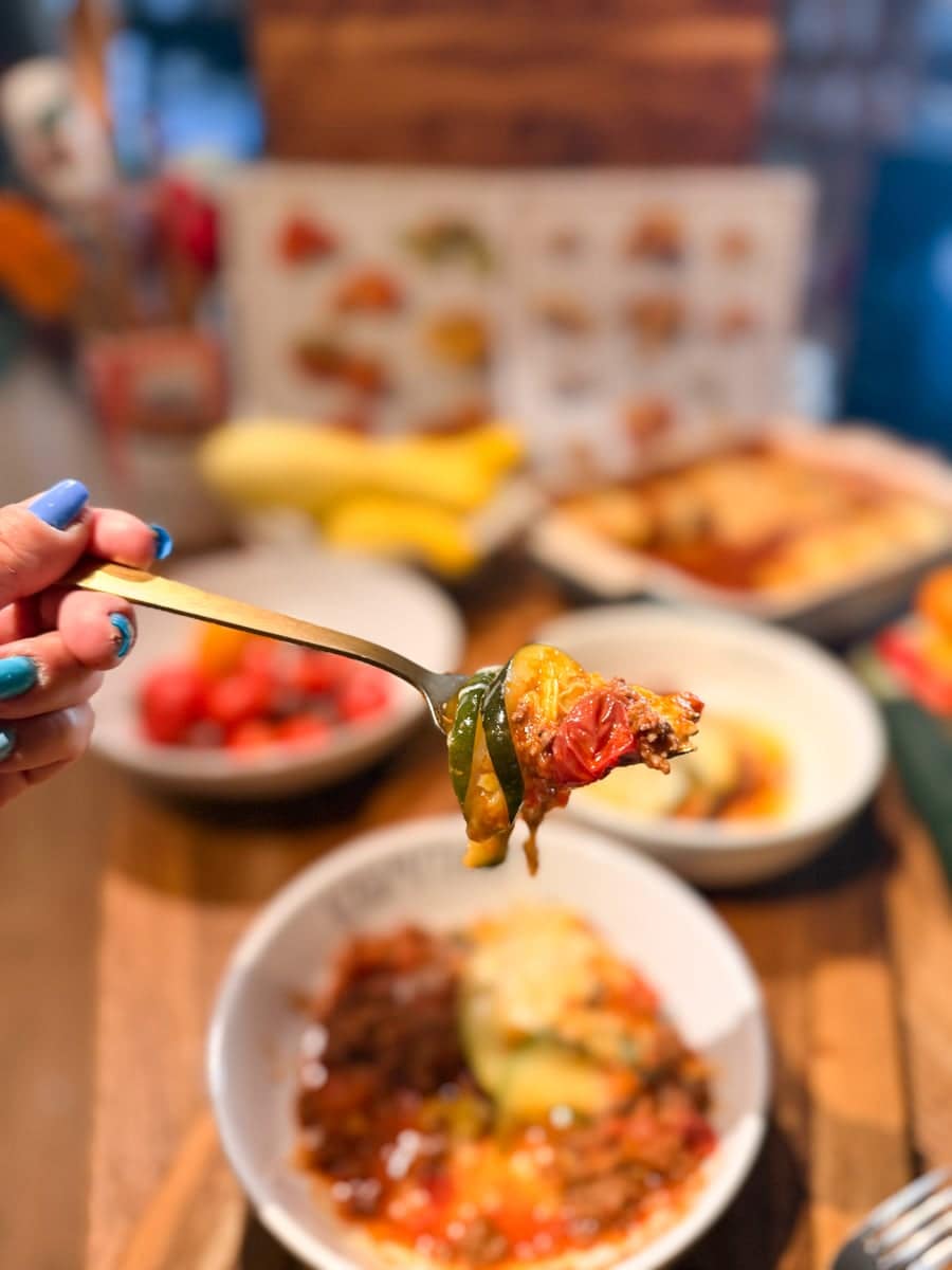 A fork holding a bite of homemade ratatouille with roasted zucchini, tomato, sage, and Parmesan cheese, with the finished dish blurred in the background.