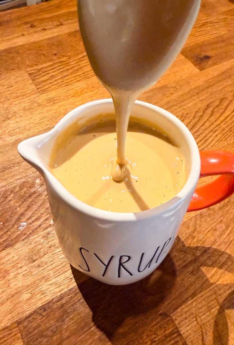 Homemade maple butter being poured into a small white syrup pitcher on a wooden kitchen counter.
