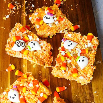 Five Halloween Rice Krispies treats topped with candy corn and white ghost decorations, arranged on a wooden serving board.