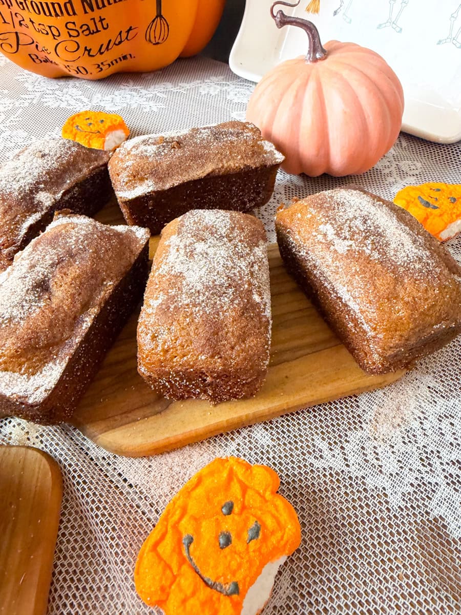 Five golden-brown pumpkin bread mini loaves dusted with cinnamon sugar on a wooden serving board, surrounded by decorative pumpkins and festive Halloween treats.