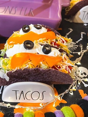 Halloween monster tacos in purple taco shells decorated with sour cream and black olive eyes, orange bell pepper smiles, and colorful spooky decorations.