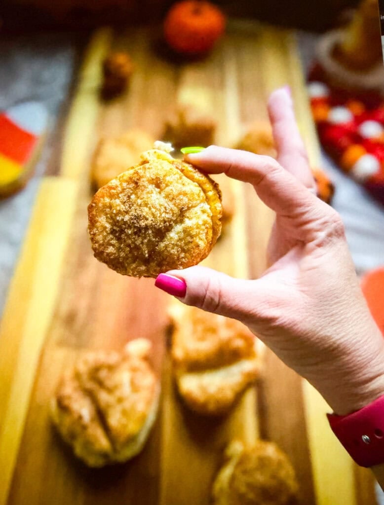 A golden brown pumpkin-shaped hand pie with a sugar-dusted puff pastry crust held above a wooden board of freshly baked pumpkin pastries