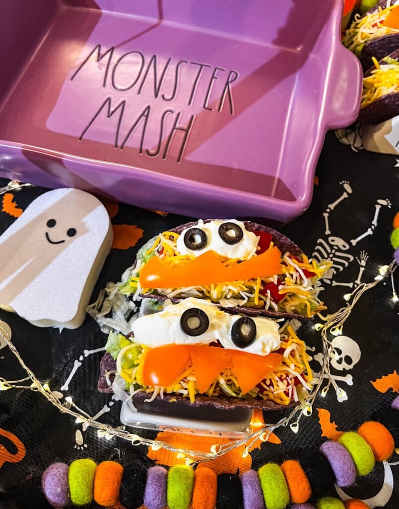Two Halloween monster tacos with purple shells, black olive eyes, and orange bell pepper smiles displayed beside a purple Monster Mash dish and ghost decoration.