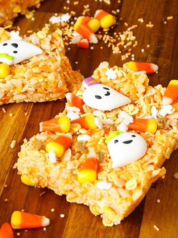 Halloween Rice Krispies Treats topped with candy corn, white ghost candies, and festive Halloween sprinkles on a wooden cutting board.