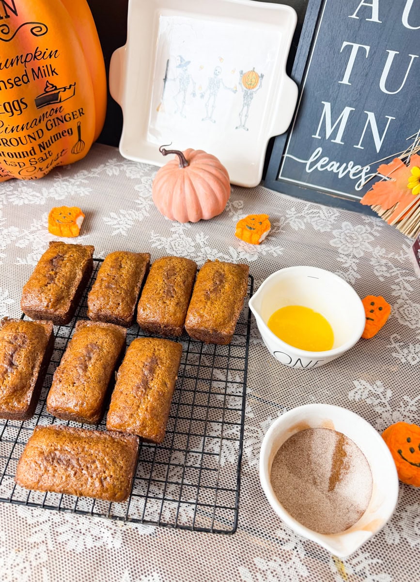 Eight golden-brown mini pumpkin bread loaves cooling on a wire rack beside bowls of melted butter and cinnamon sugar.