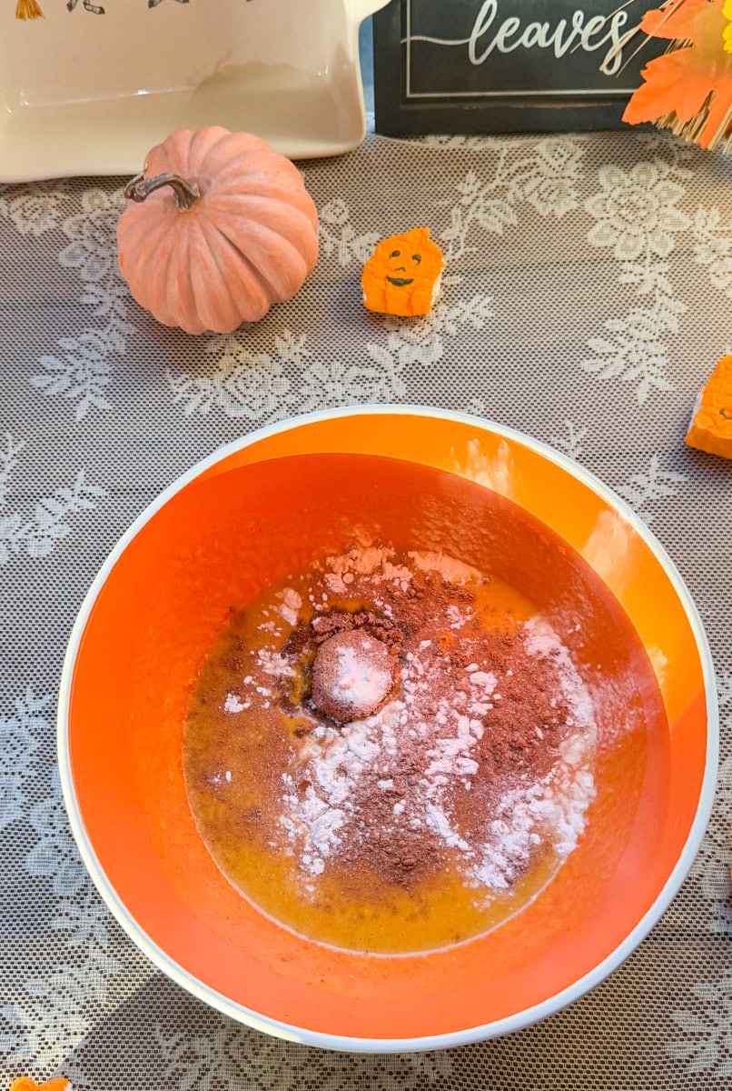 Ground cinnamon and pumpkin spice added to an orange mixing bowl of wet ingredients for homemade mini pumpkin bread loaves.
