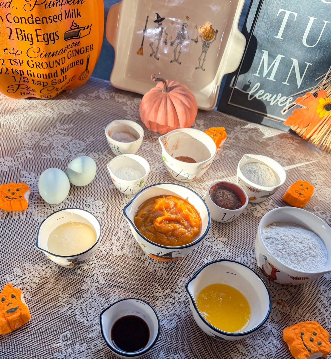 Ingredients for homemade mini pumpkin bread loaves with pumpkin puree, sweetened condensed milk, flour, melted butter, eggs, vanilla extract, cinnamon, pumpkin pie spice, sugar, baking soda, and baking powder arranged on a fall-themed table.