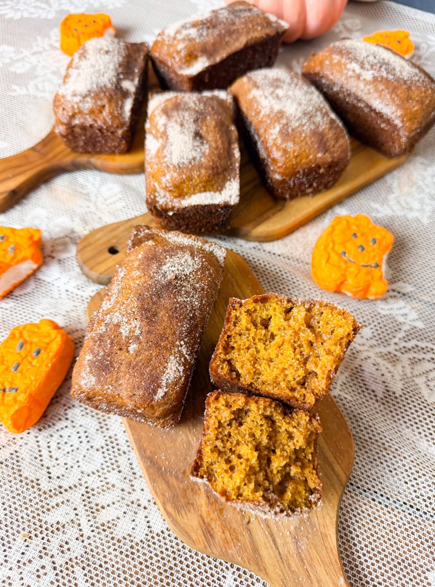 Mini pumpkin bread loaves with cinnamon sugar crust on wooden serving boards, featuring a sliced loaf showing the moist pumpkin crumb and festive Halloween decorations.