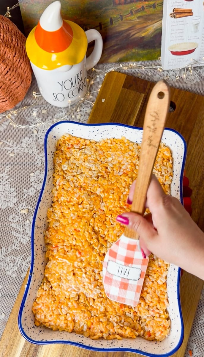 Using a spatula to gently press the candy corn Rice Krispies mixture into an even layer in a prepared baking dish for homemade Halloween treats.
