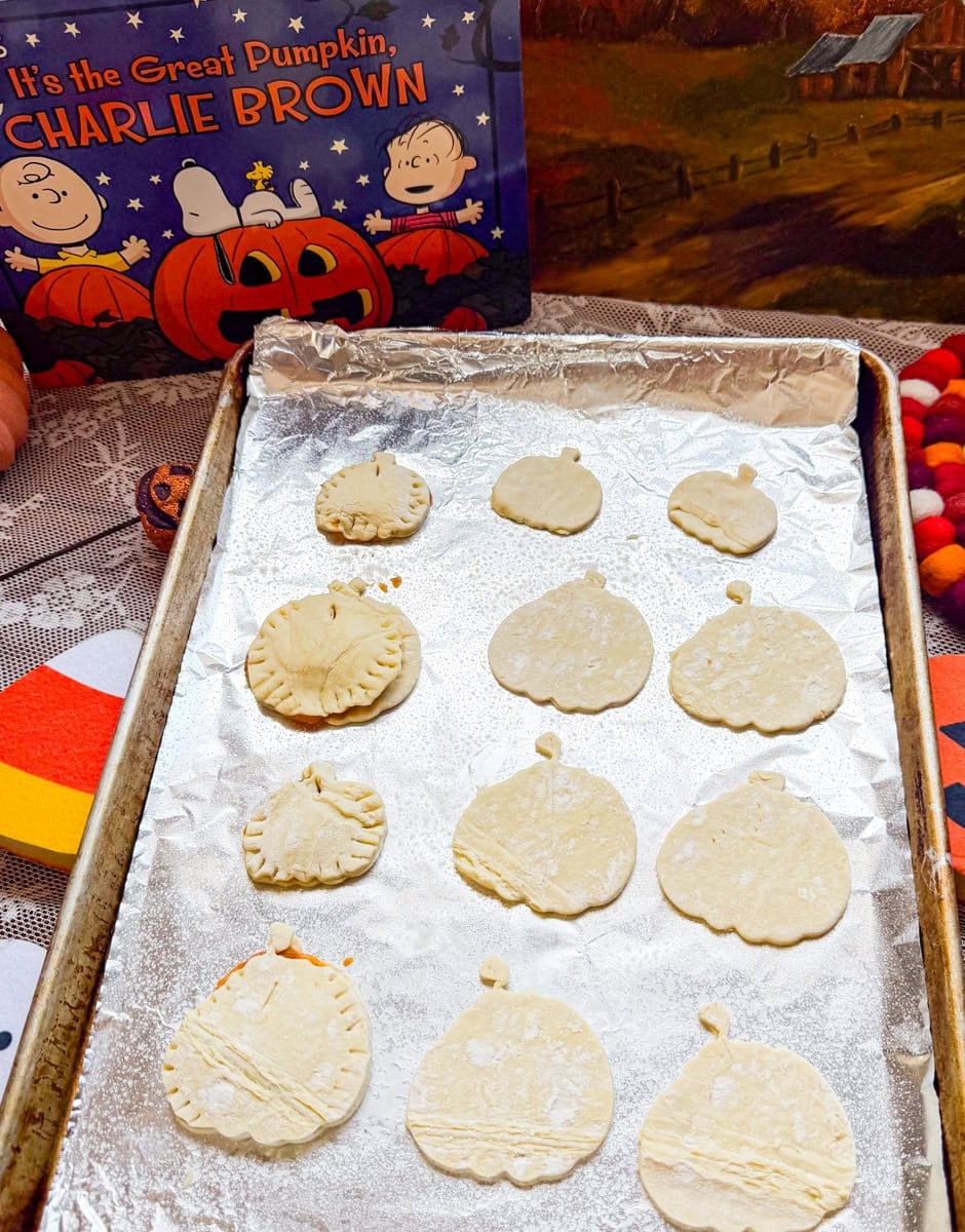 Unbaked pumpkin-shaped puff pastry hand pies arranged on a foil-lined baking sheet, with some edges sealed using a fork.