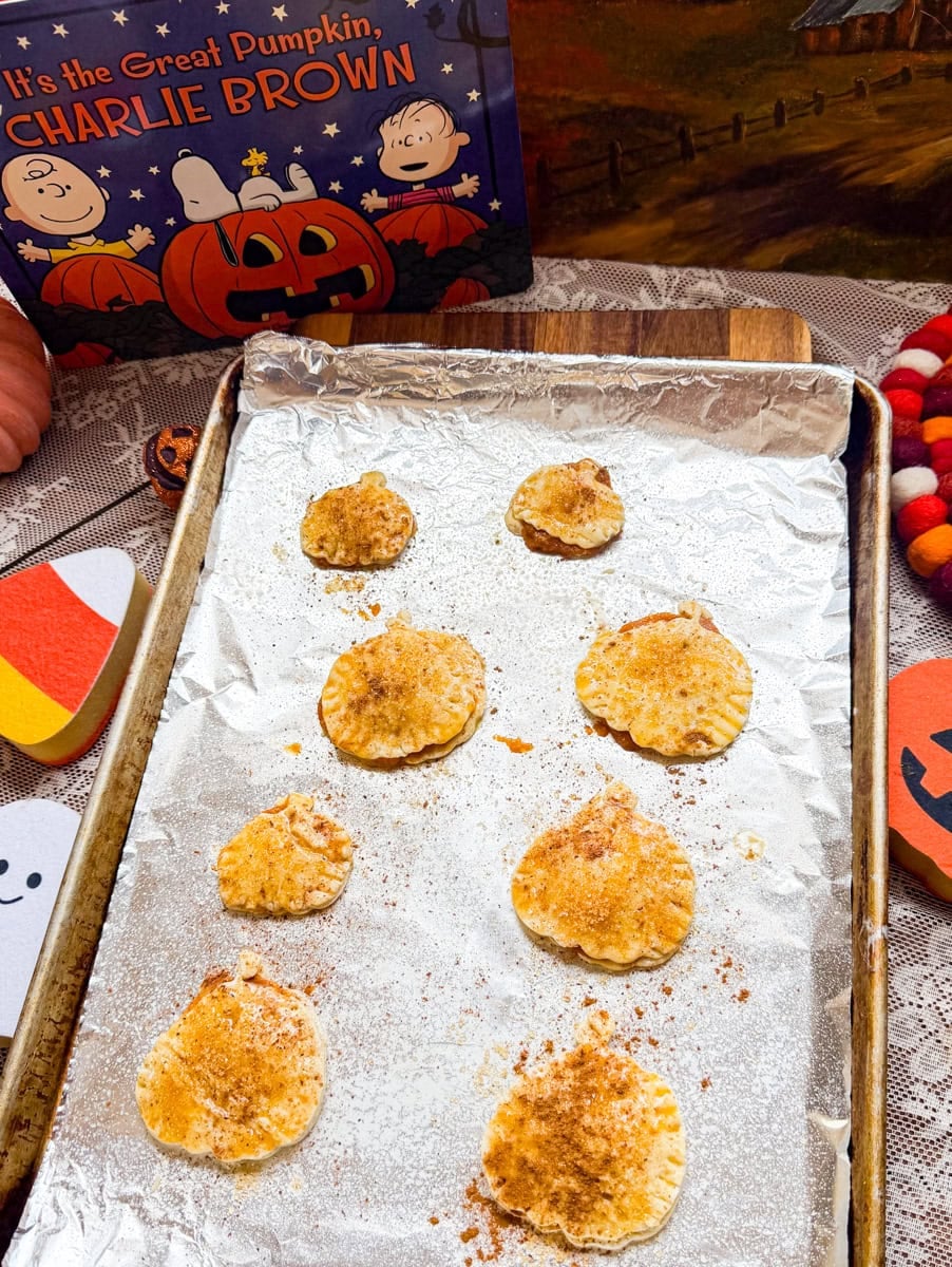Pumpkin-shaped hand pies brushed with egg wash and sprinkled with cinnamon sugar on a foil-lined baking sheet before baking.