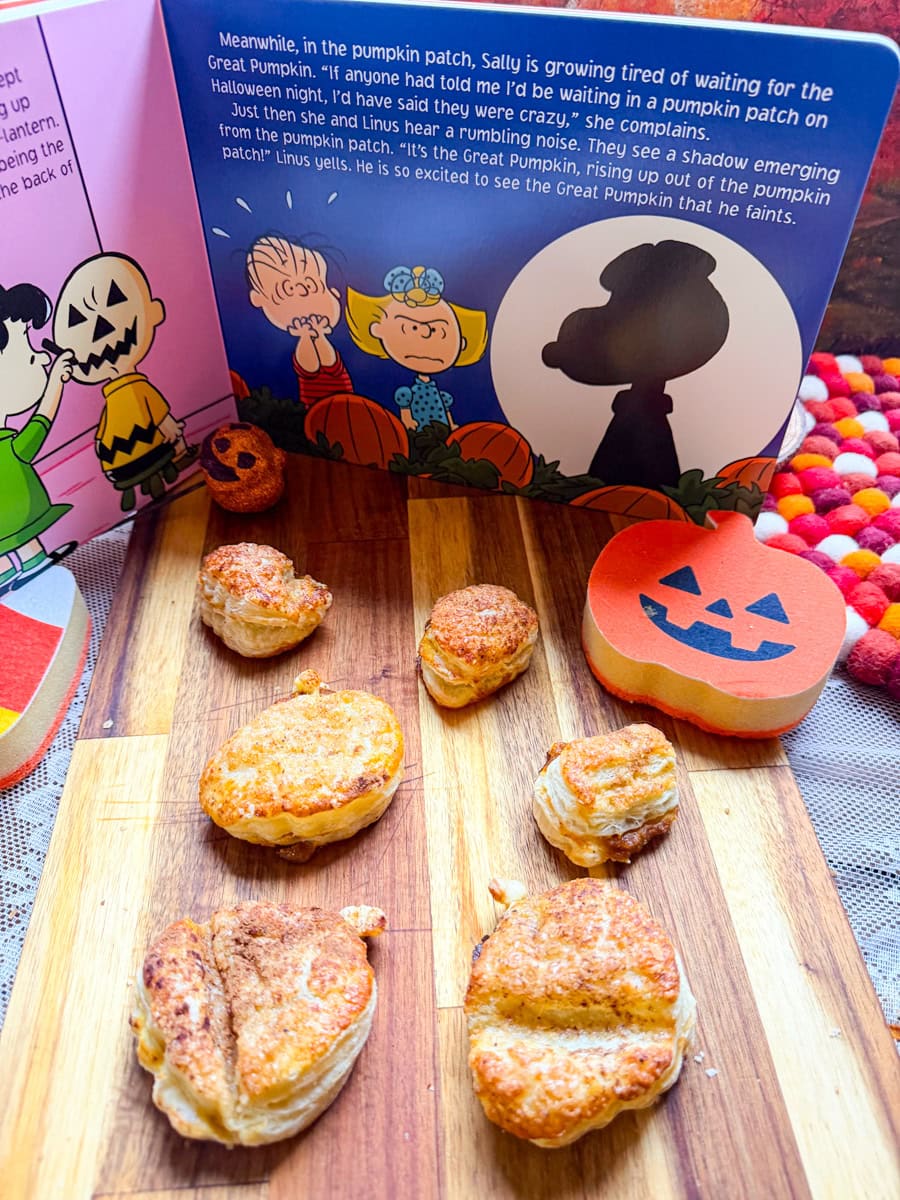 Golden brown pumpkin pie hand pies made with flaky puff pastry, arranged on a wooden cutting board with festive Halloween decorations and a Charlie Brown Great Pumpkin book.
