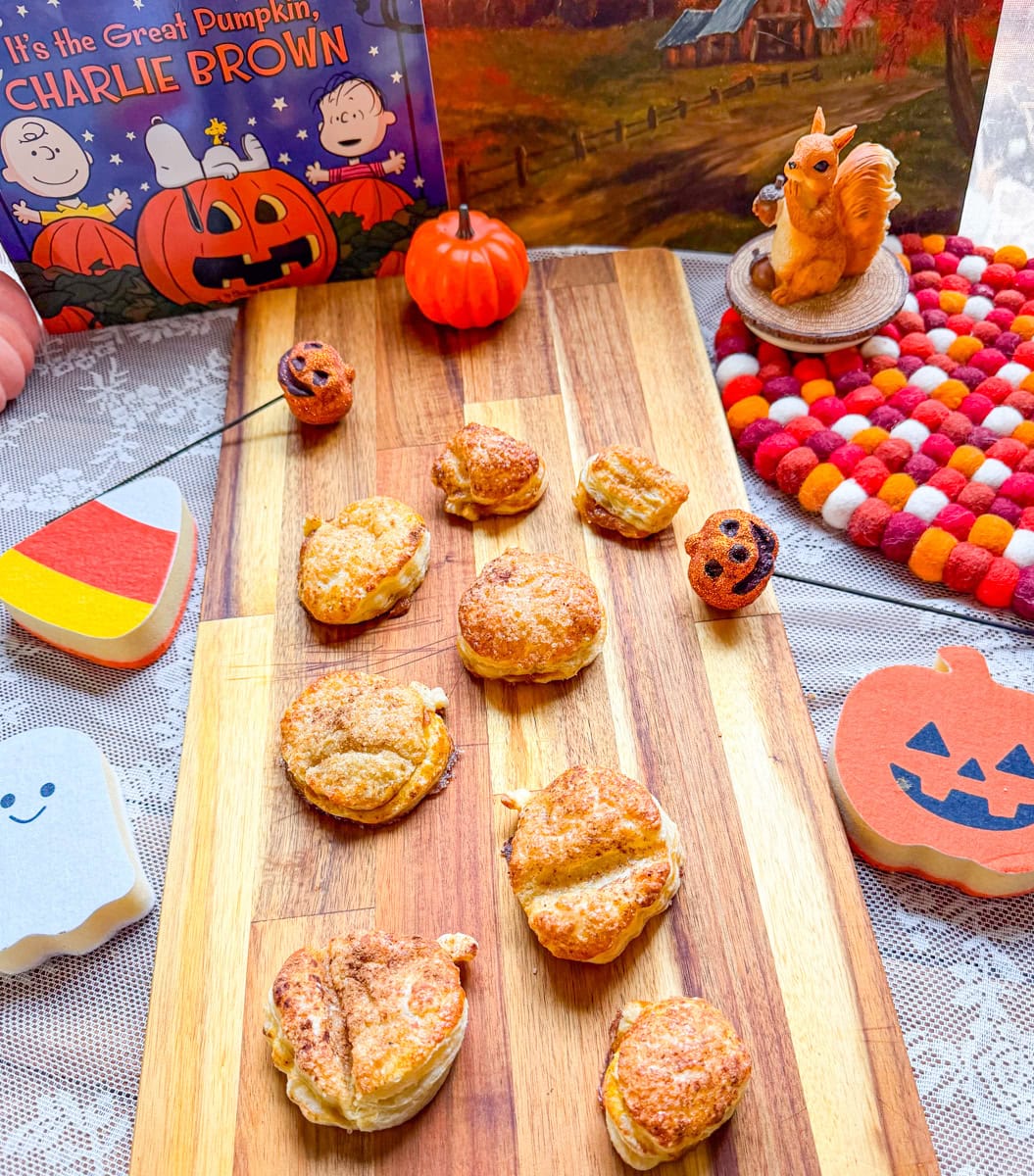 Golden brown pumpkin pie hand pies made with flaky puff pastry, arranged on a wooden cutting board with festive Halloween decorations, candy corn accents, and a Charlie Brown Great Pumpkin book.
